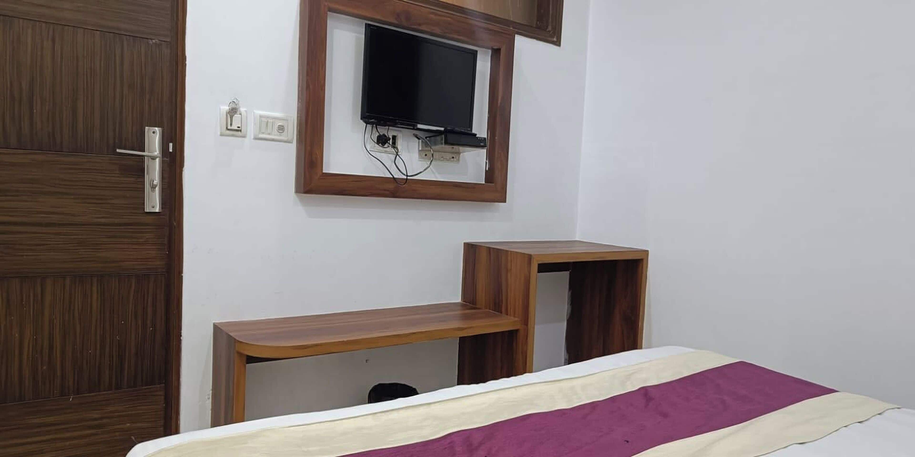 A bedroom with a wooden door, a TV mounted on the wall, and a bed with a purple and white blanket.