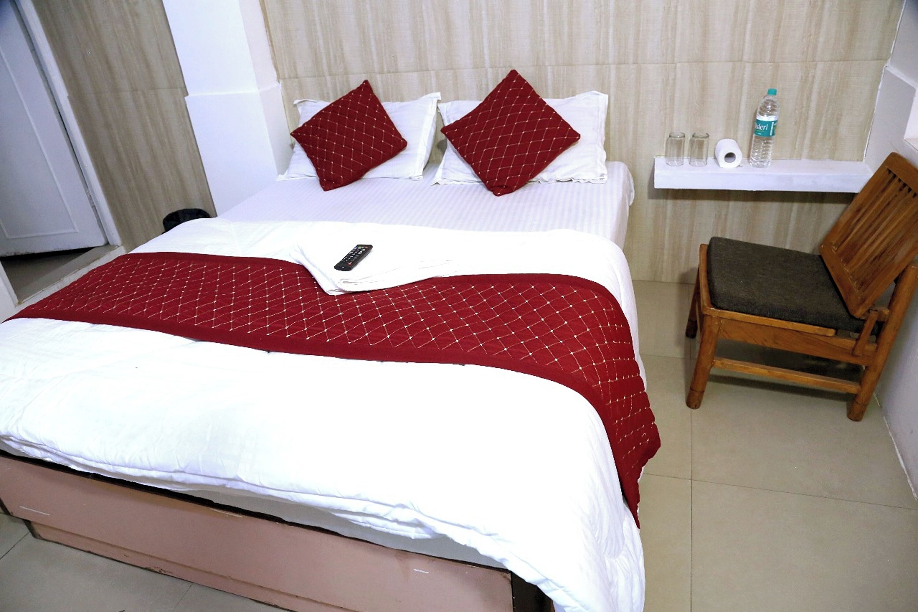 A hotel room with a bed, a chair, a shelf with toiletries, and a remote control on the bed.