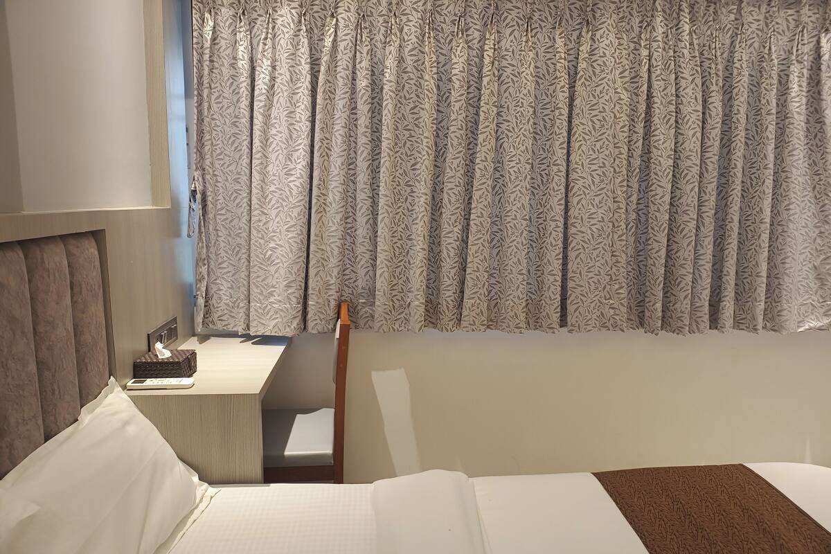 A hotel room with a bed, a bedside table, a desk, and a window with patterned curtains.