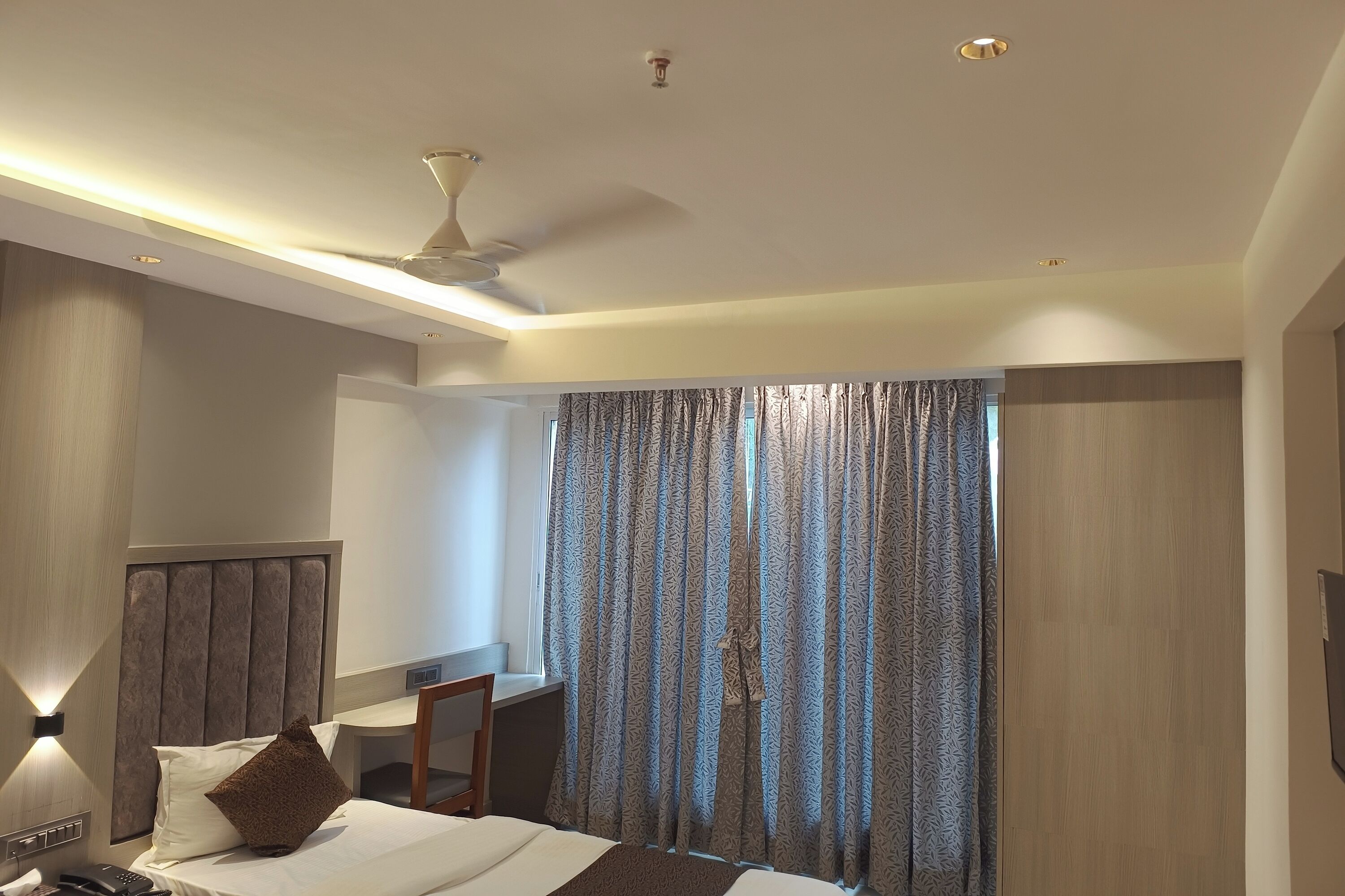 A hotel room with a bed, a desk, and a chair. There is a window with curtains, a ceiling fan, and recessed lighting.