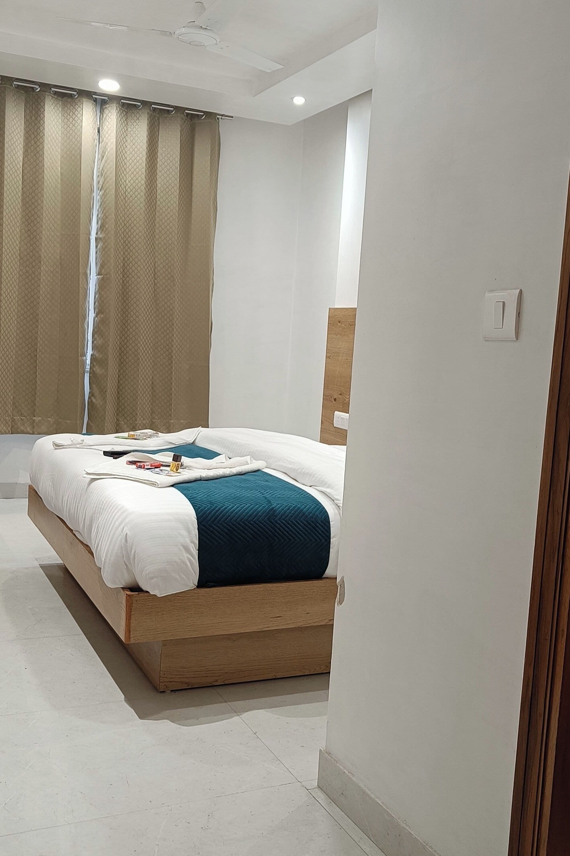 A modern bedroom with a wooden bed, white bedding, a teal throw, and a window with beige curtains.