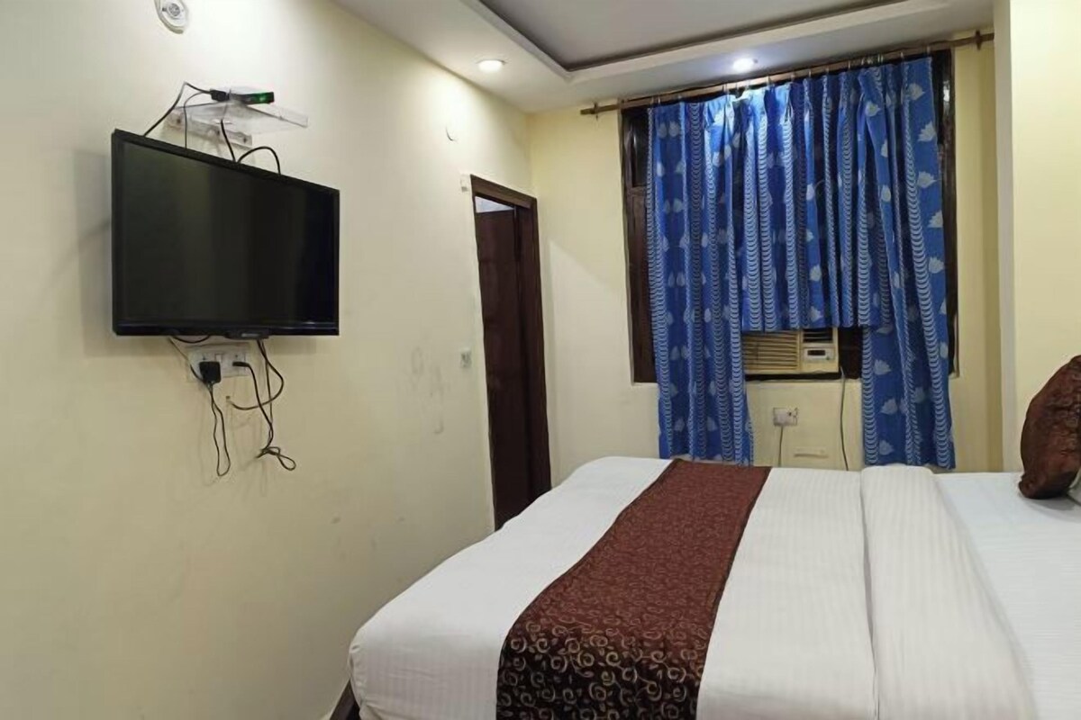 A hotel room with a bed, a television mounted on the wall, a window with curtains, and an air conditioning unit.