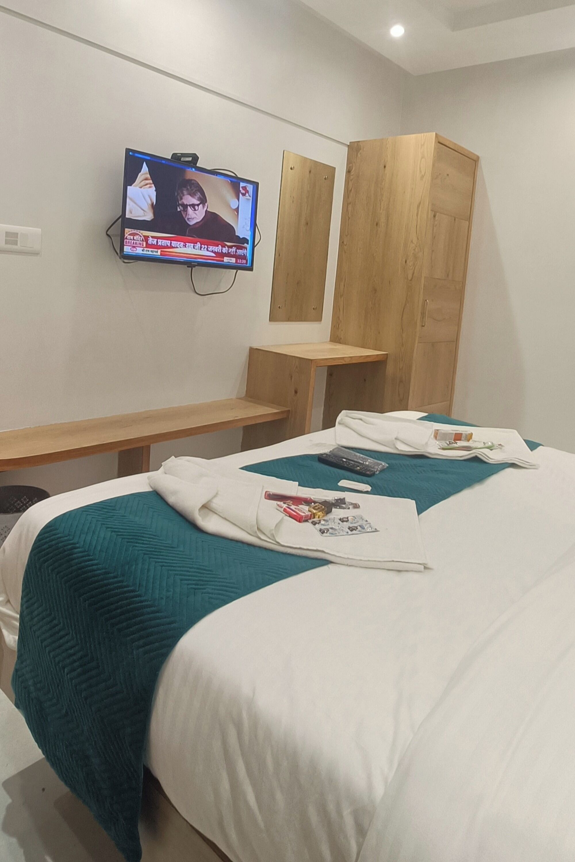 A hotel room with a bed, a TV mounted on the wall, a wooden wardrobe, and a small wooden table.