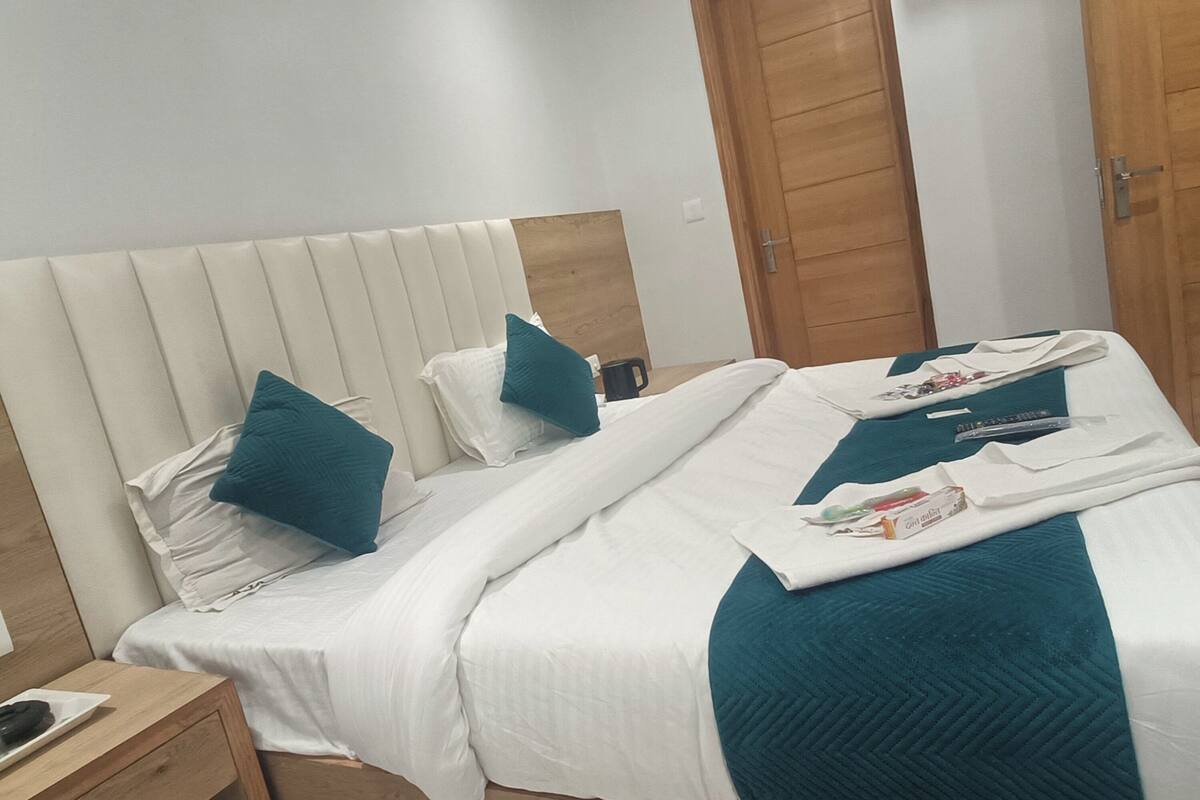 Un lit bien fait, avec une literie blanche et des touches de couleur turquoise, une table de chevet en bois avec une télécommande, et une armoire en bois en arrière-plan.
