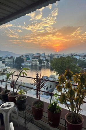Un balcon agrémenté de plantes en pot, offrant une vue sur un paysage urbain avec des bâtiments et une rivière, au coucher du soleil.