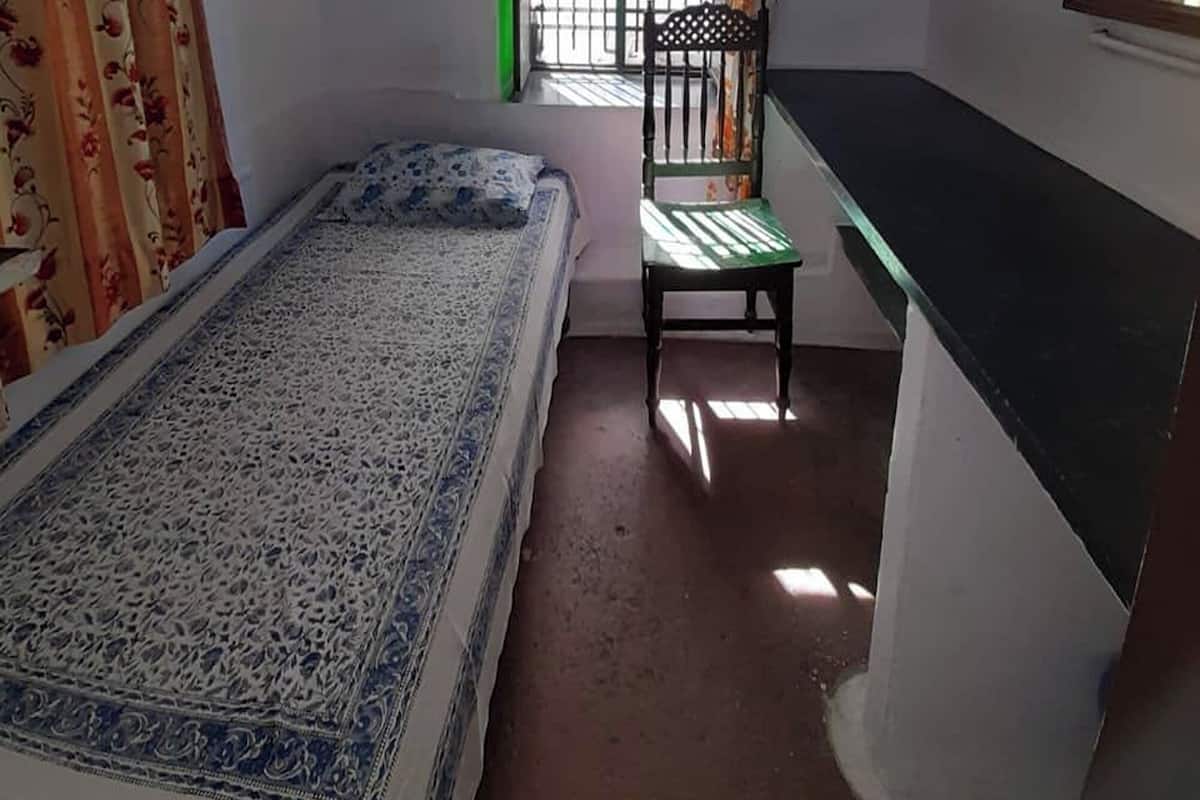 A small, single-room bedroom with a bed, a chair, a desk, and a ceiling fan.