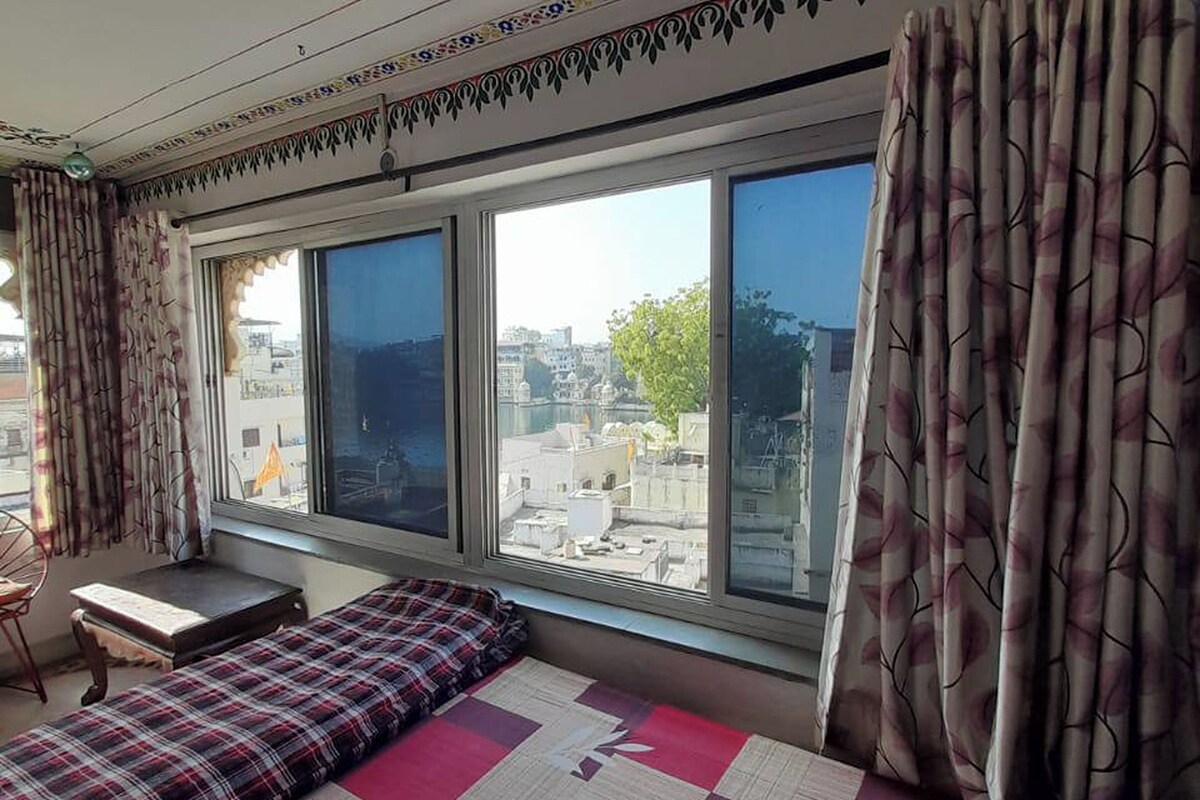 A room with a large window, a bed with a checkered bedspread, a small table, and a chair.