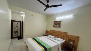 A bedroom with a bed, wooden headboard, bedside tables, a ceiling fan, and a painting on the wall. - Savitri Bungalow (Jaoli)