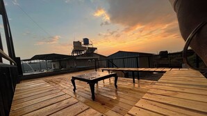 A wooden deck with tables and chairs, overlooking a cityscape at sunset. - Savitri Bungalow (Jaoli)