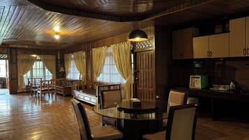 A spacious room with a wooden ceiling, a round glass table, and wooden chairs. There are curtains on the windows, and a vintage radio on a table.