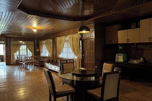 Una habitación amplia con techo de madera, una mesa redonda de vidrio y sillas de madera. Hay cortinas en las ventanas y una radio antigua sobre una mesa.