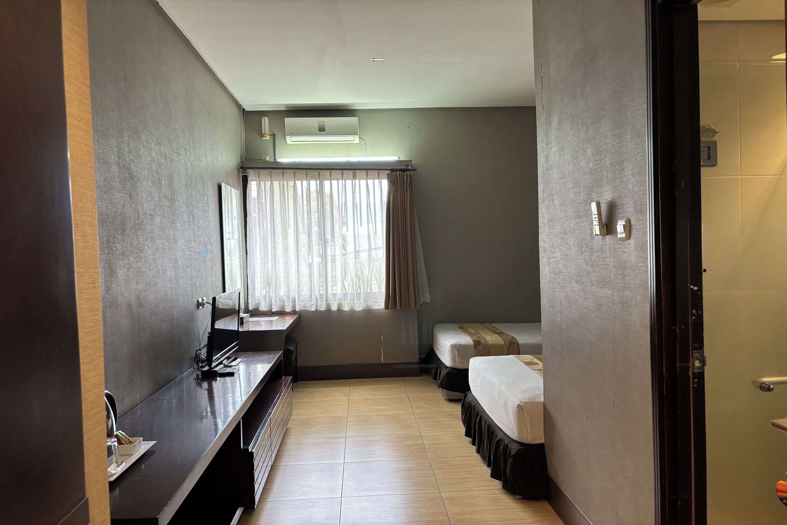 Habitación de hotel con un tocador largo de madera oscura, cama con colchón blanco y falda oscura, ventana con cortinas de gasa y aire acondicionado de pared.