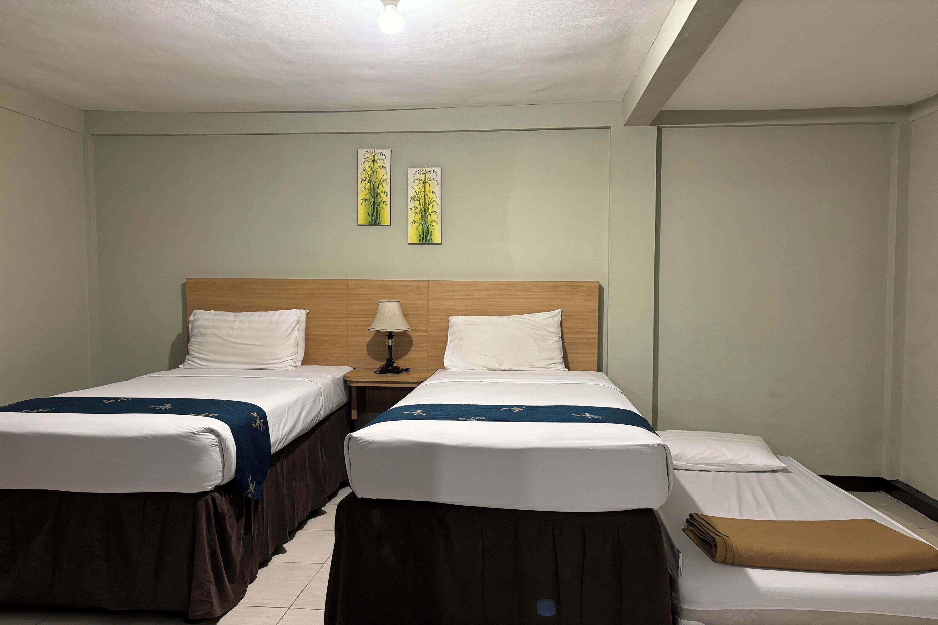 A hotel room with two single beds, a wooden headboard, a nightstand with a lamp, and two wall art pieces.