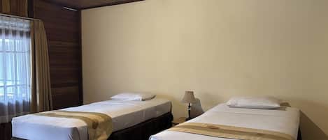 A room with two beds, each with a white mattress and a brown bed skirt. There is a window with curtains on the left, and a lamp on the right side of the room.