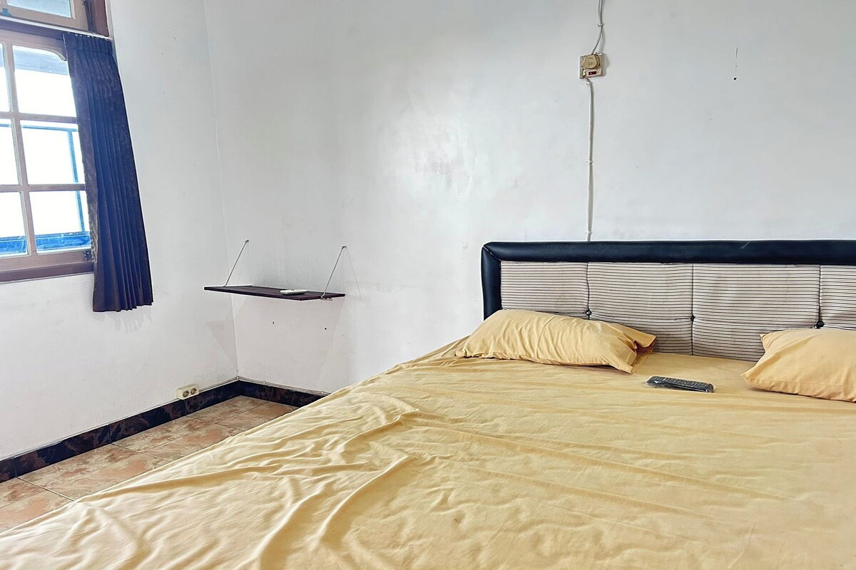 A room with a bed, a window with curtains, a wall-mounted shelf, and a power outlet.