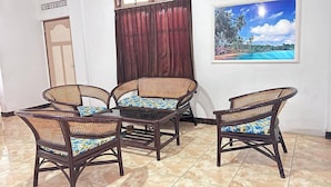 Une pièce comprenant une table à manger et quatre chaises en osier, une grande fenêtre avec des rideaux et, au mur, un tableau représentant une plage tropicale.