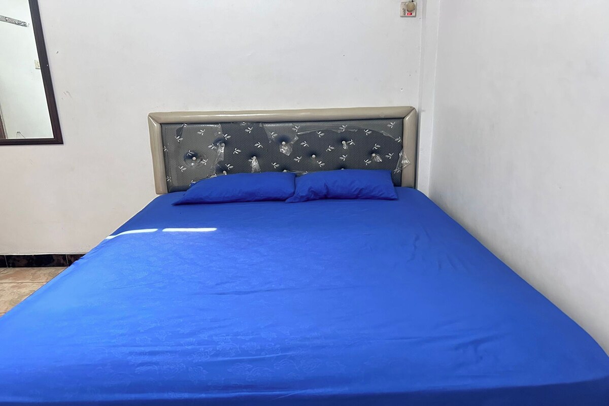 Uma cama de solteiro com colchão e almofadas azuis combinando, cabeceira com padrão decorativo e parede lisa com um pequeno interruptor de luz.
