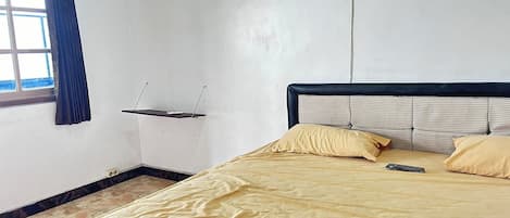 Una habitación con cama, ventana con cortinas, estante montado en la pared y toma de corriente.