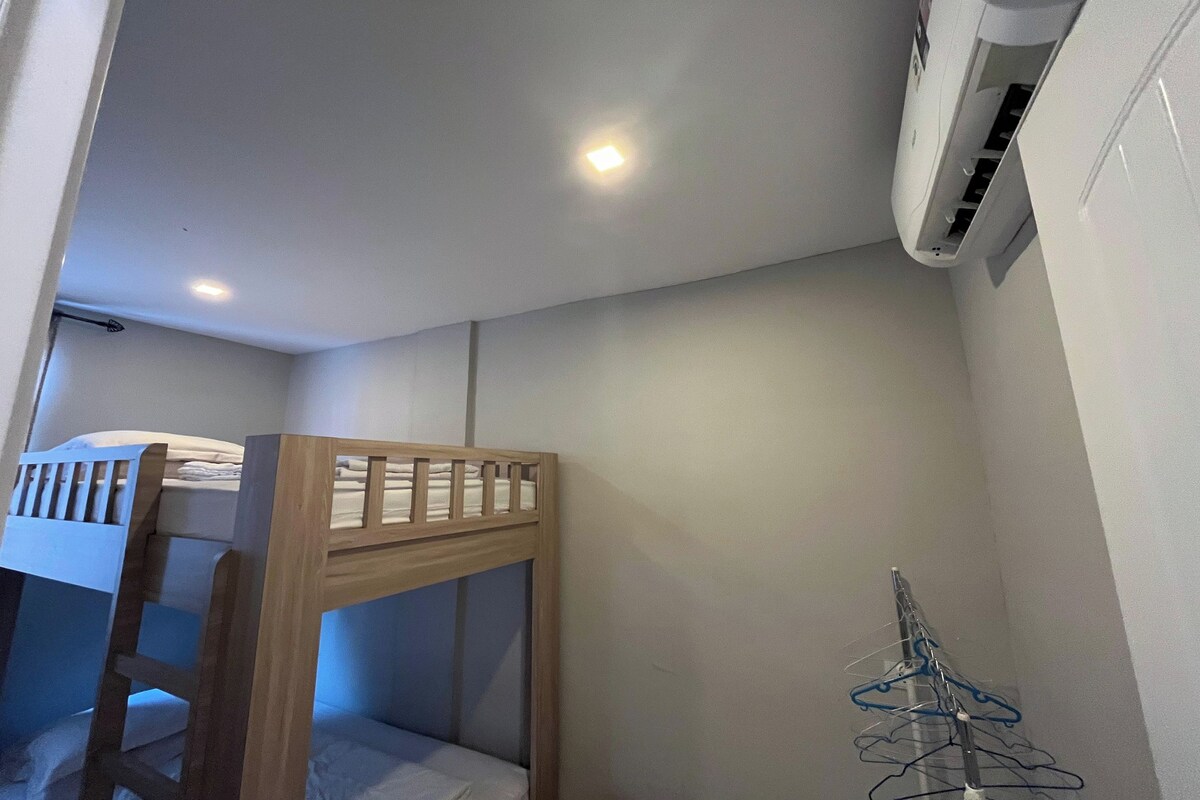 Un lit superposé avec une structure en bois et un matelas blanc, un climatiseur fixé au mur et un étendoir à linge.