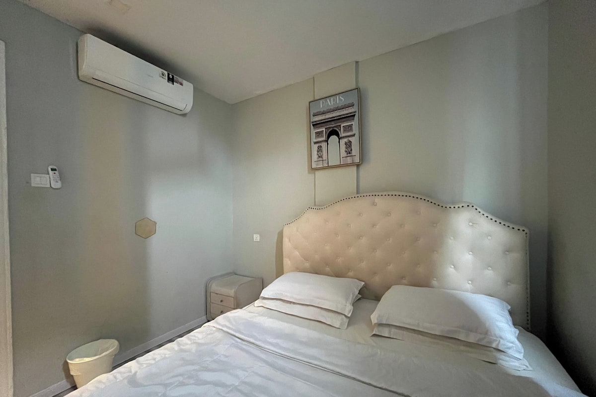A bedroom with a large bed, a nightstand, a bedside table, an air conditioning unit, and a framed picture on the wall.