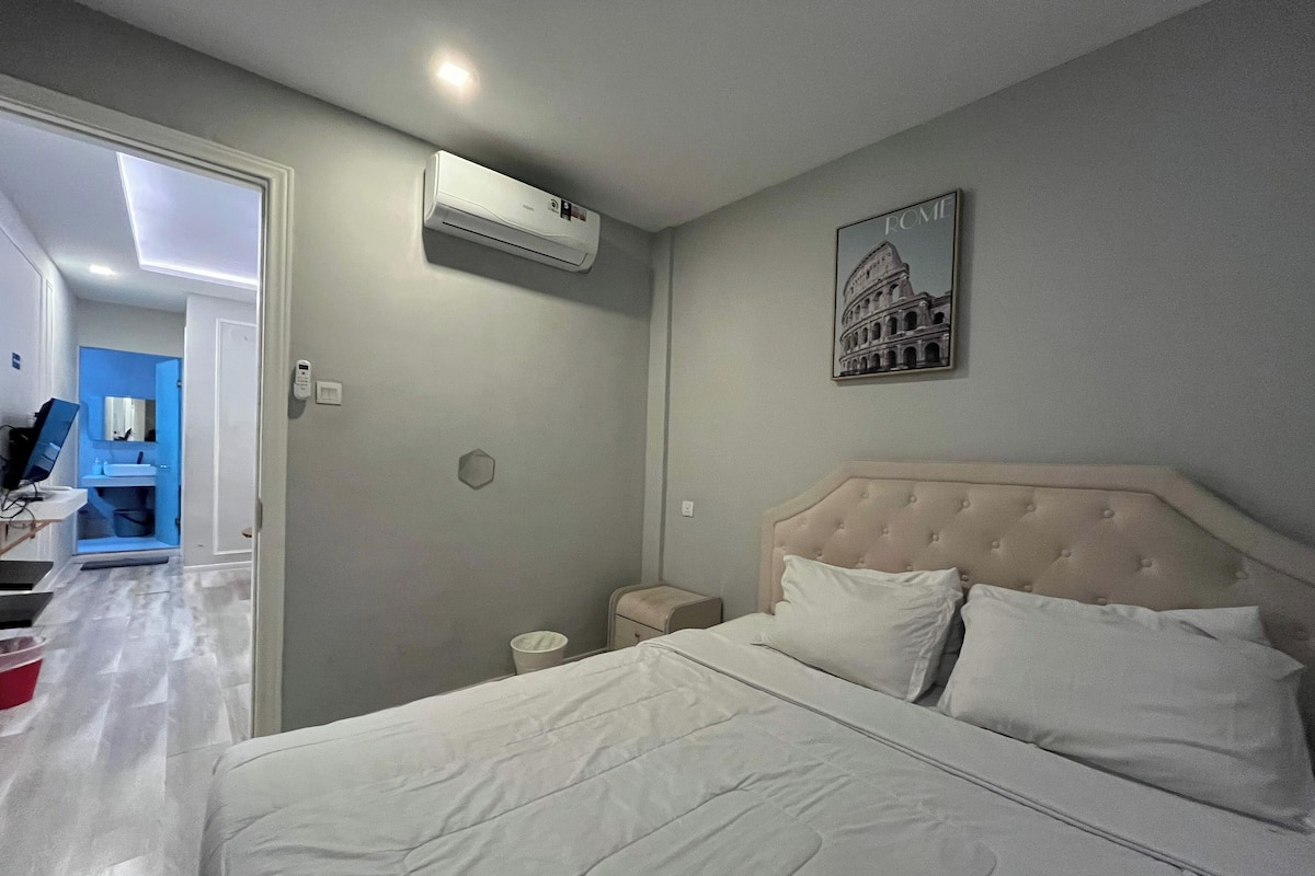A hotel room with a bed, a nightstand, a wall-mounted air conditioner, a bathroom with a mirror and sink, and a framed picture of the Colosseum.