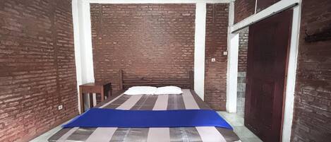 A room with a bed, a small table, and a chair. The walls are made of brick, and there is a wooden door.