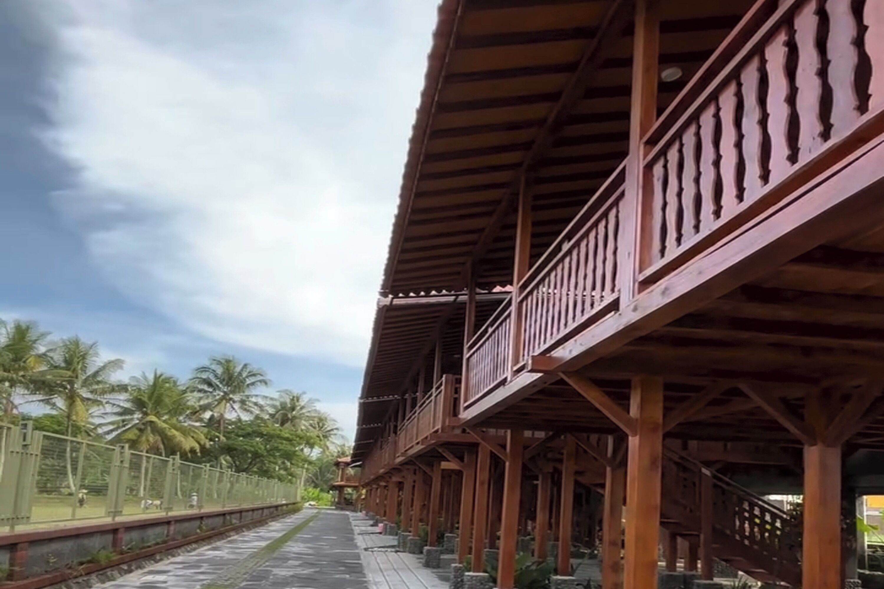 A wooden structure with balconies, a paved walkway, and palm trees in the background.