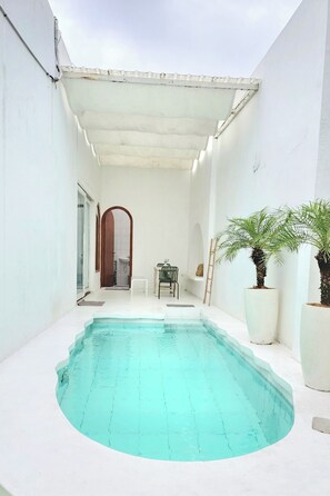A small, enclosed swimming pool area with a white interior, a door leading to a bathroom, and two potted palm trees. - Floraison Living Villa Yogyakarta (Ngaglik)