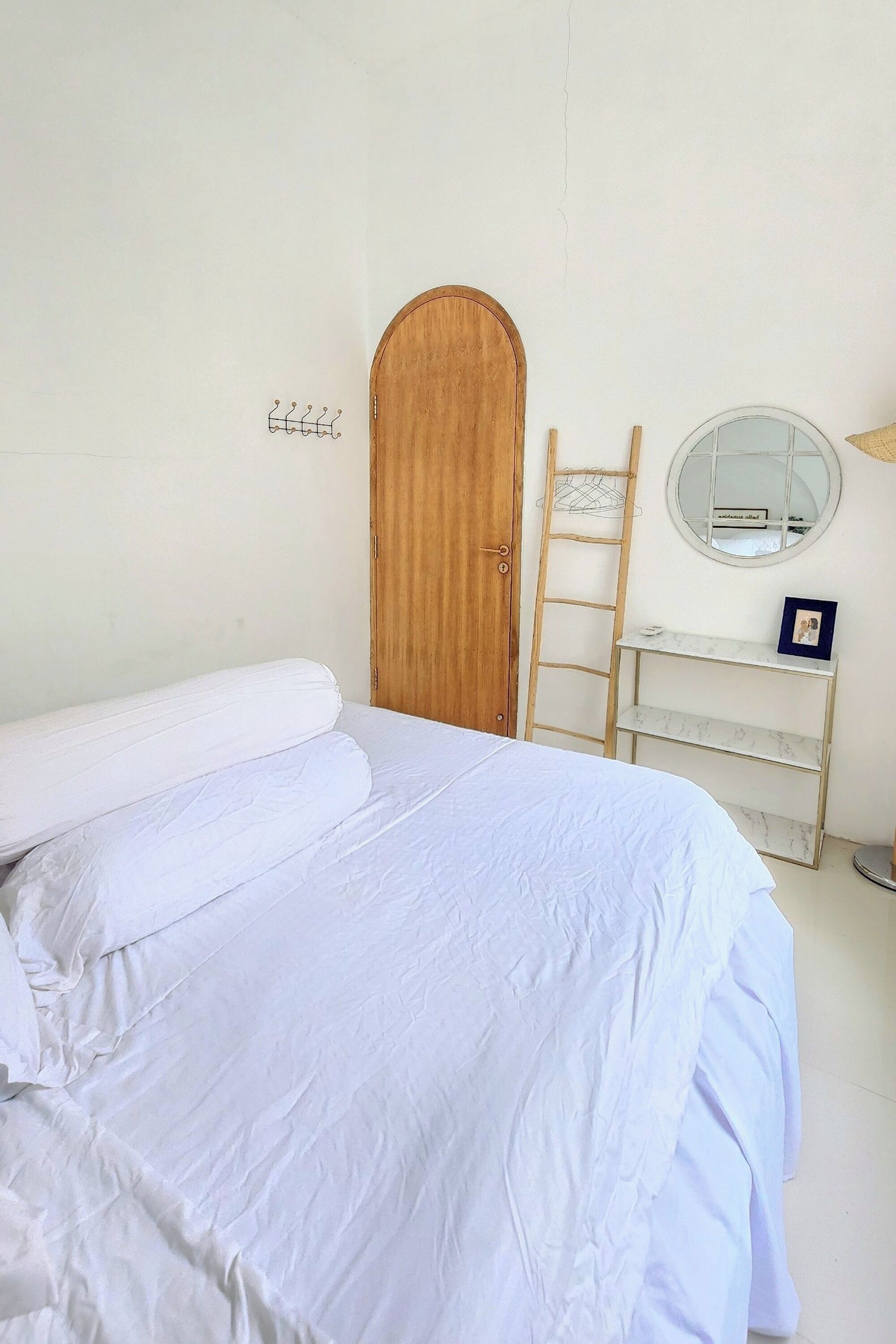 A neatly made bed with white linens, a wooden door, a ladder, a small shelf with a mirror, and a framed picture on the wall.