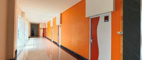 A long, well-lit hallway with orange and white walls, a shiny tiled floor, and doors marked with room numbers.