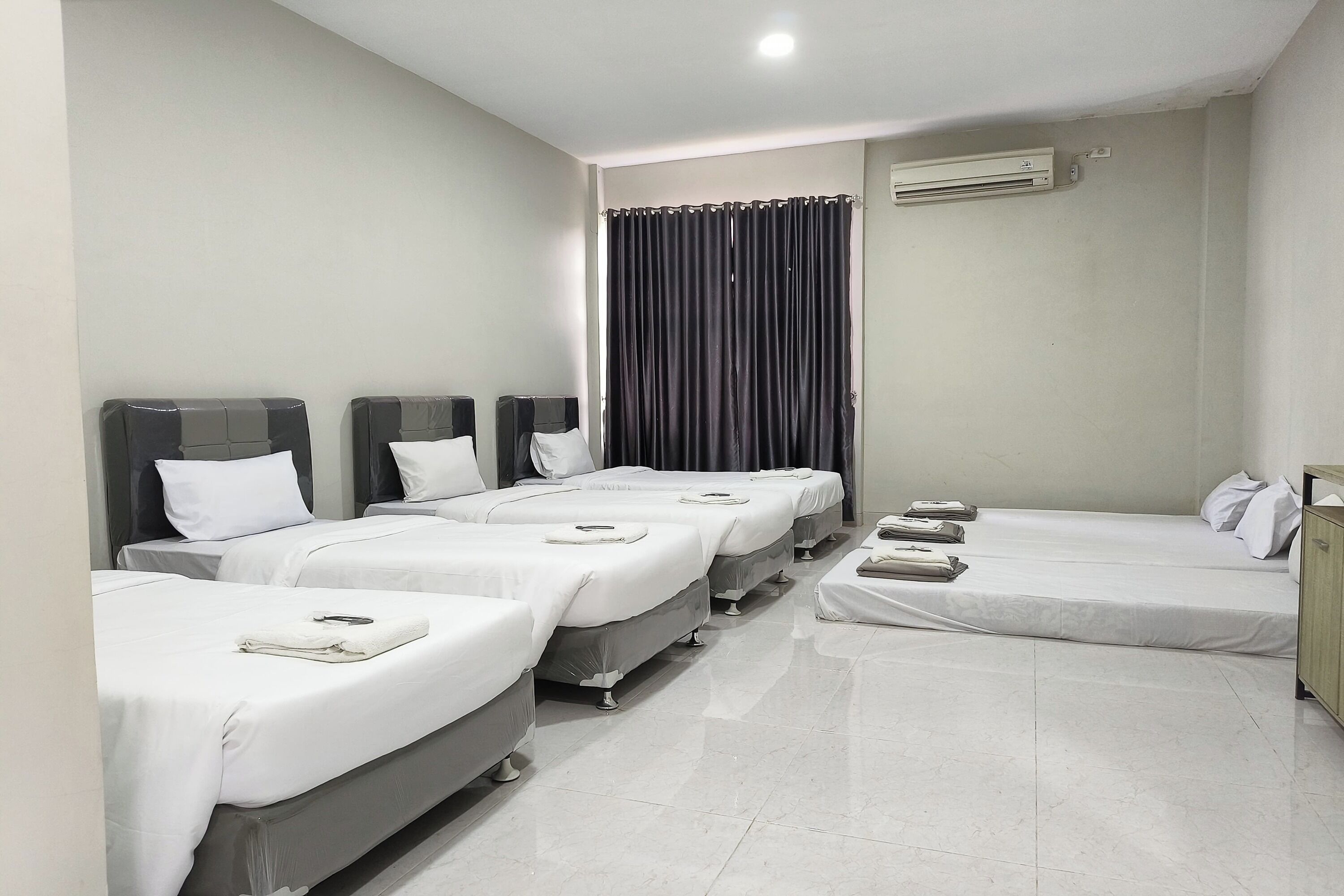 Una habitación de hotel con cuatro camas individuales, un ventanal con cortinas oscuras, un armario de madera y un aire acondicionado instalado en la pared.