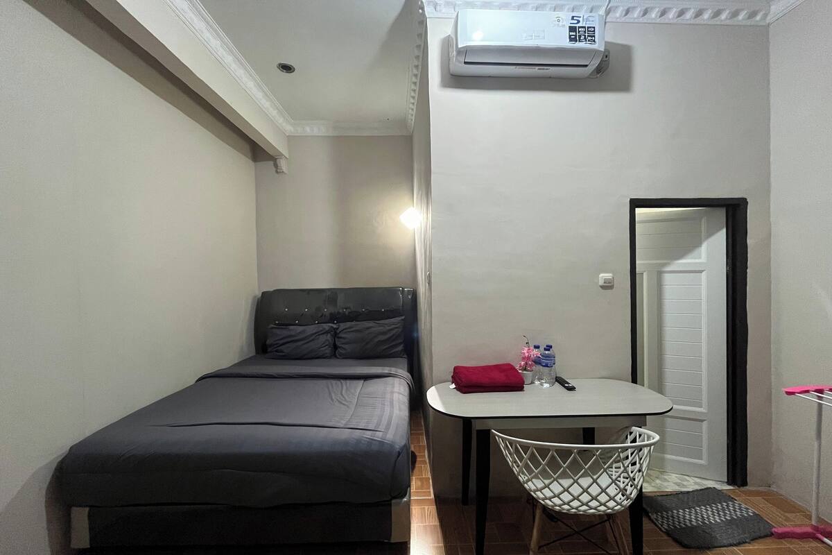 A small, compact room with a single bed, a small table with a chair, an air conditioning unit, and a door.