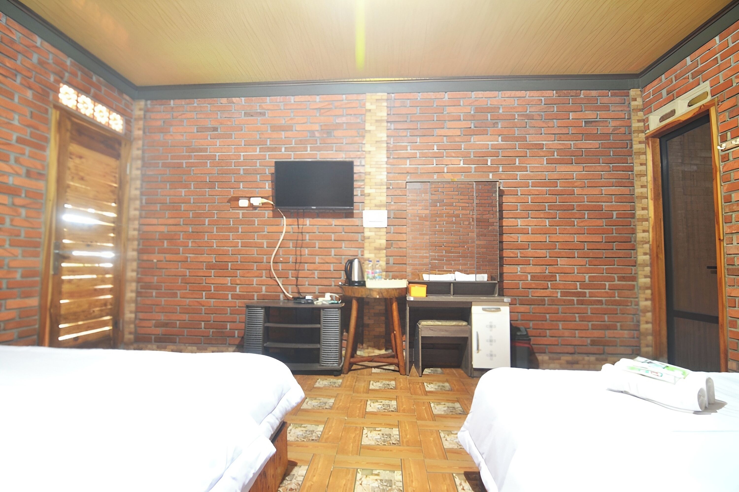 Una habitación con dos camas, un televisor instalado en la pared de ladrillo, un escritorio con una silla y un refrigerador pequeño.