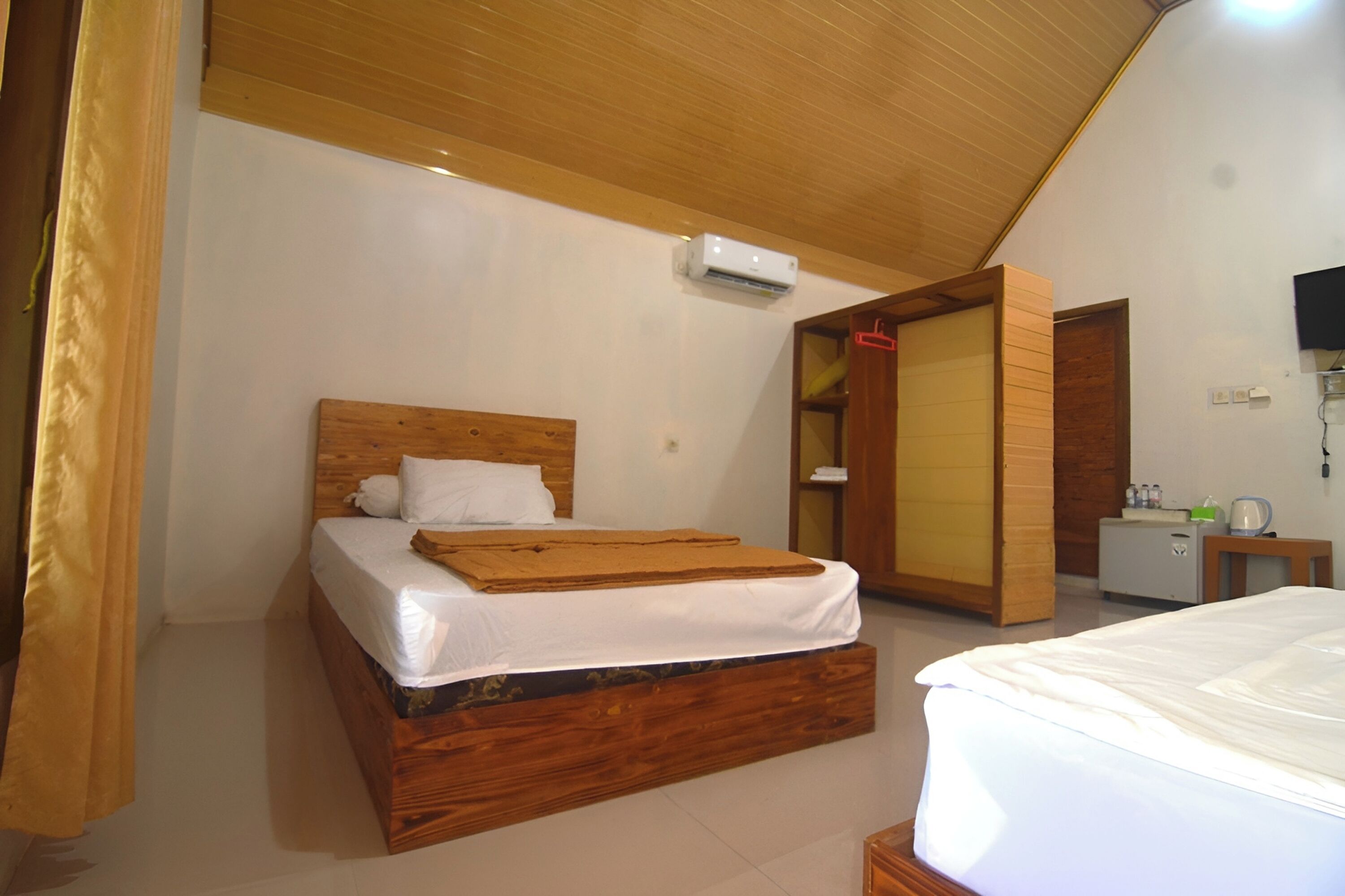 Un dormitorio con cama de madera, ropa de cama blanca, un equipo de aire acondicionado, una mesita de noche pequeña y un armario.