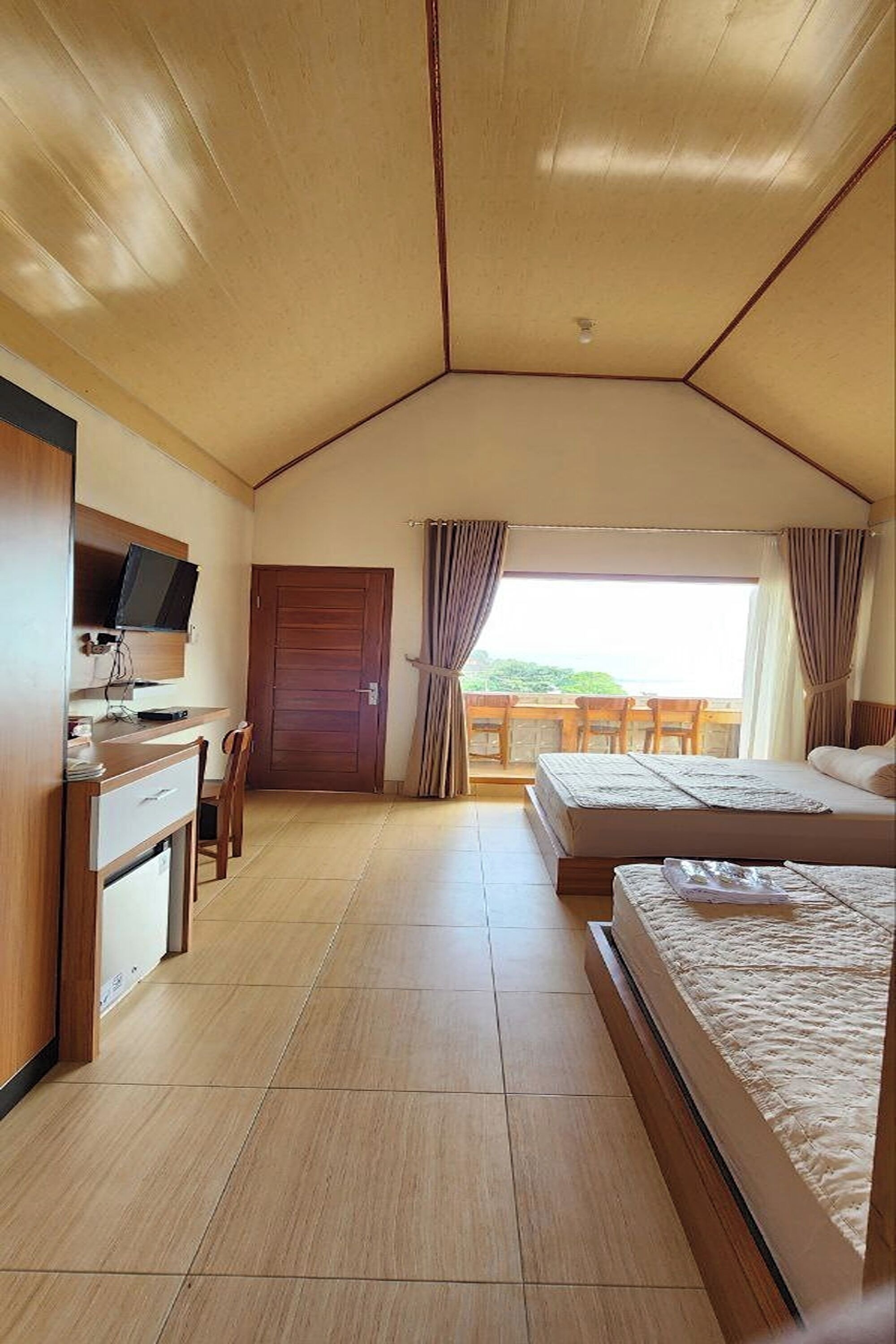 A room with a wooden ceiling, a desk with a television, two beds, and a balcony with a view.