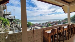 Un balcon avec une table et des chaises en bois, donnant sur une ville côtière et l’océan.