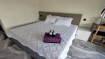A neatly made bed with a white quilted bedspread and pillows, a small white nightstand with a cup and a decorative mirror, and a wooden chair.