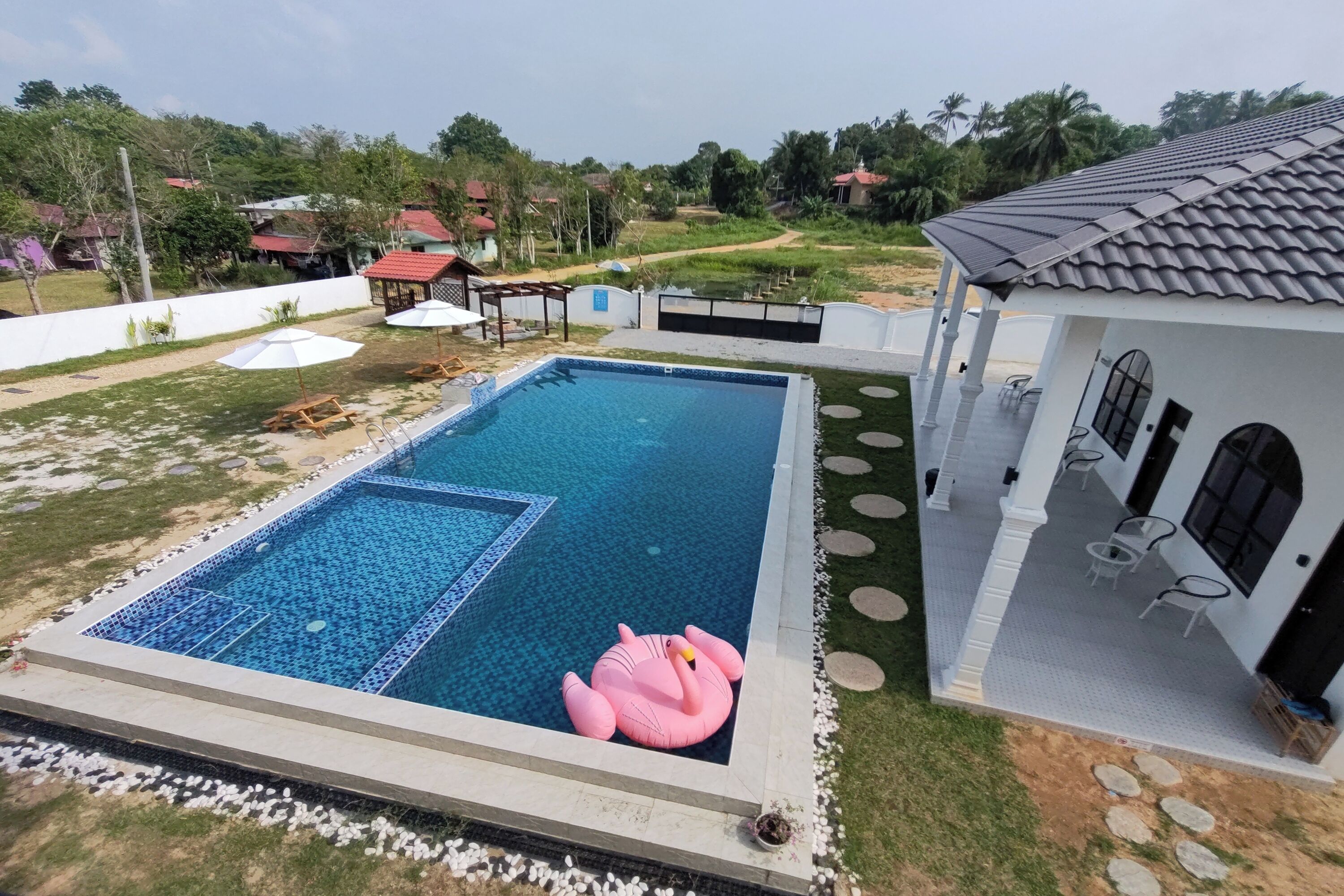 A swimming pool with a pink flamingo float, surrounded by a white building with outdoor seating and a view of a green landscape.