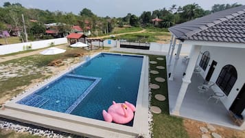 A swimming pool with a pink flamingo float, surrounded by a white building with outdoor seating and a view of a green landscape.