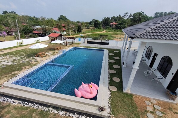 Una piscina con un flotador de flamenco rosa, rodeada por un edificio blanco con área de estar al aire libre y vistas a un paisaje verde.