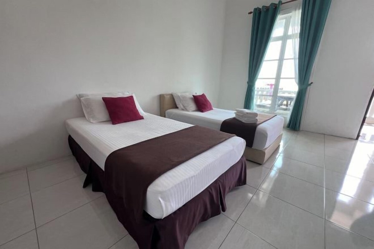 Una habitación de hotel con dos camas, cada una con colchón blanco y falda marrón. Hay una ventana con cortinas color aguamarina, y el piso es de baldosas.