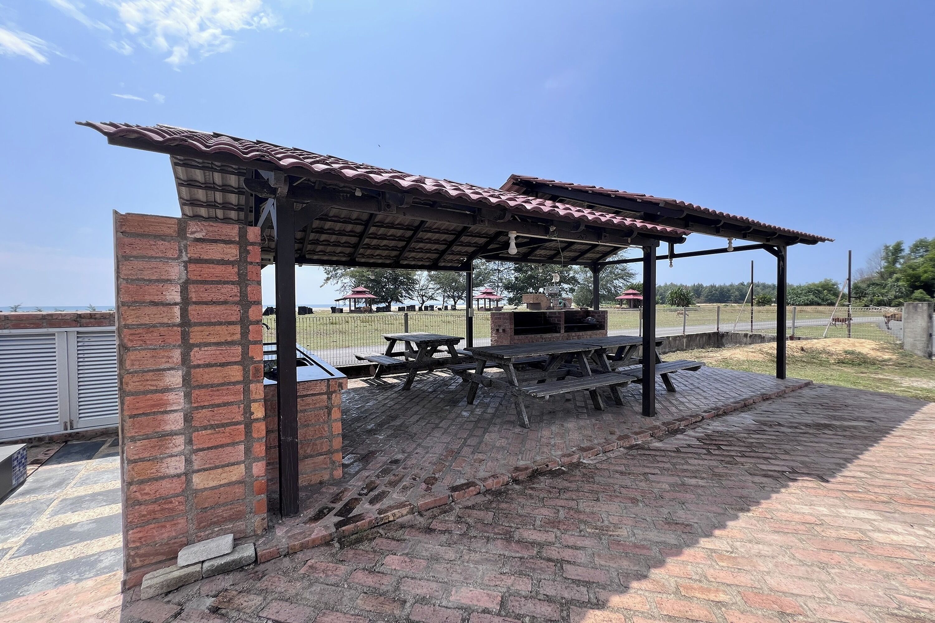 A covered outdoor seating area with brick walls and a tiled roof, featuring picnic tables and a barbecue grill.