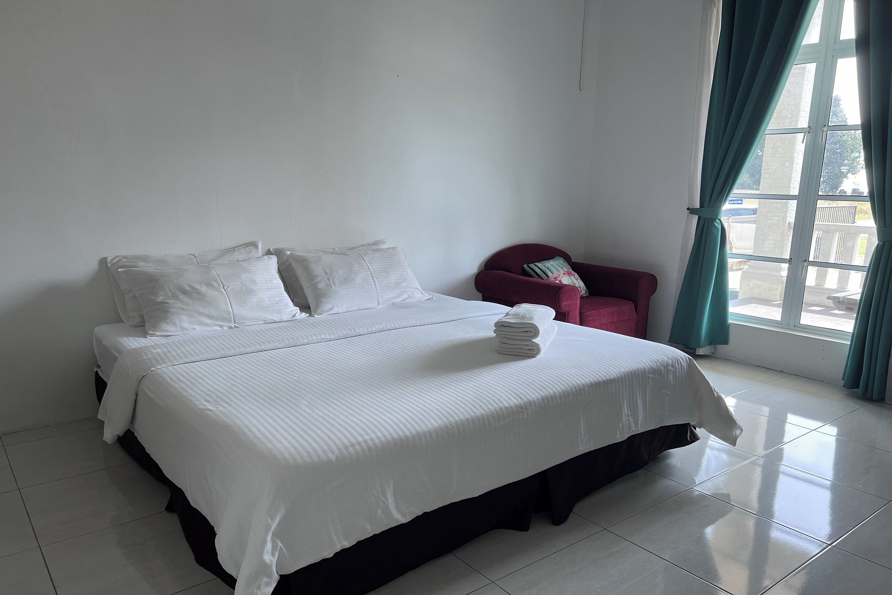 Una cama bien hecha con sábanas blancas, una silla roja y vistas a través de una ventana con cortinas.