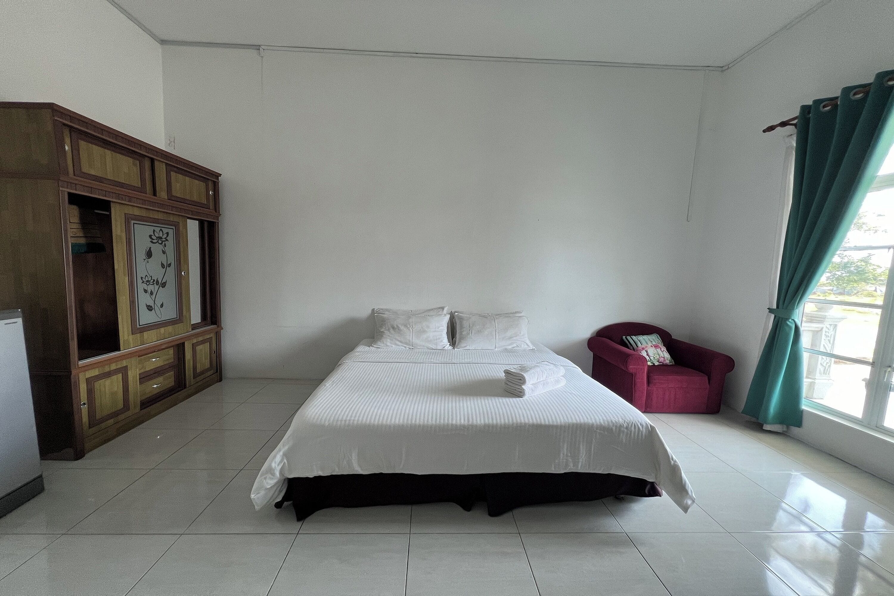 A neatly made bed in a room with a wooden cabinet and a red chair.