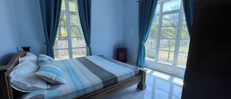Un dormitorio con cama de madera, cortinas azules y vista a palmeras a través de la ventana.