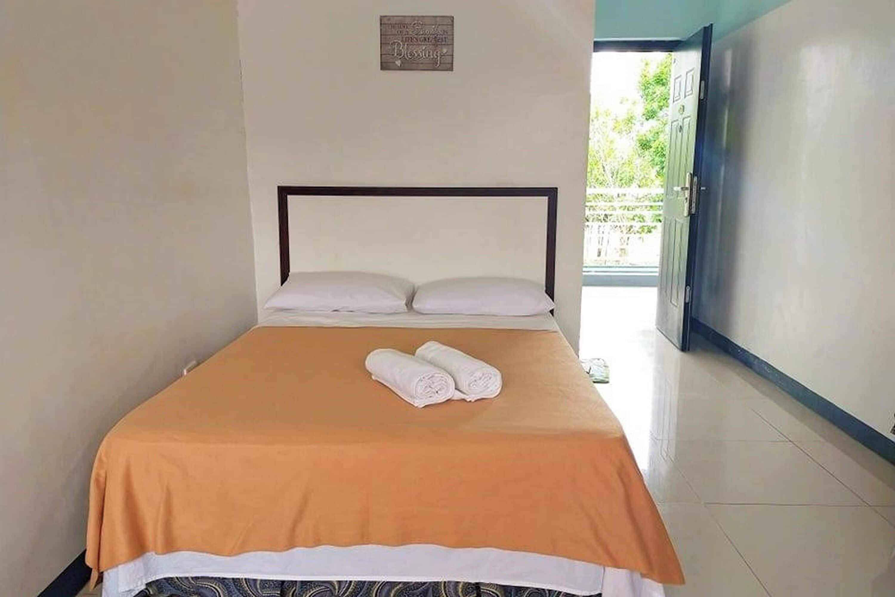 A neatly made bed with a brown bedspread and white pillows, a pair of rolled towels on the bed, and a small sign on the wall above the bed.