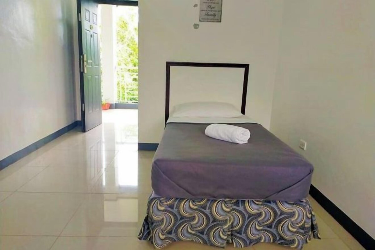 A single bed with a patterned skirt, a pillow, and a folded towel on the bed.