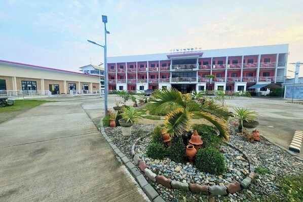 A building with a red and white facade, multiple balconies, and a landscaped garden with palm trees and potted plants. - EC Hotel near Laoag Airport (Laoag)