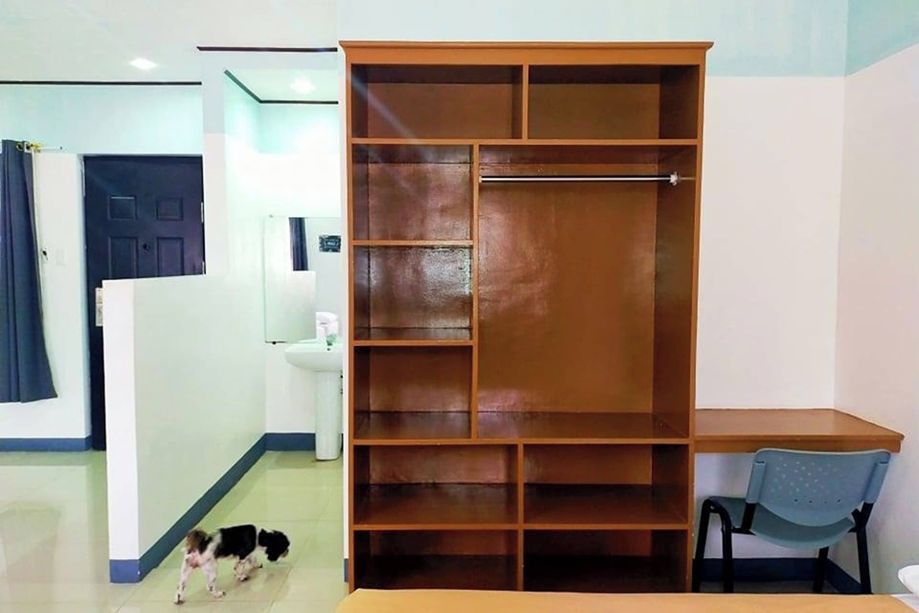 A room with a wooden wardrobe, a desk, a chair, and a small dog.