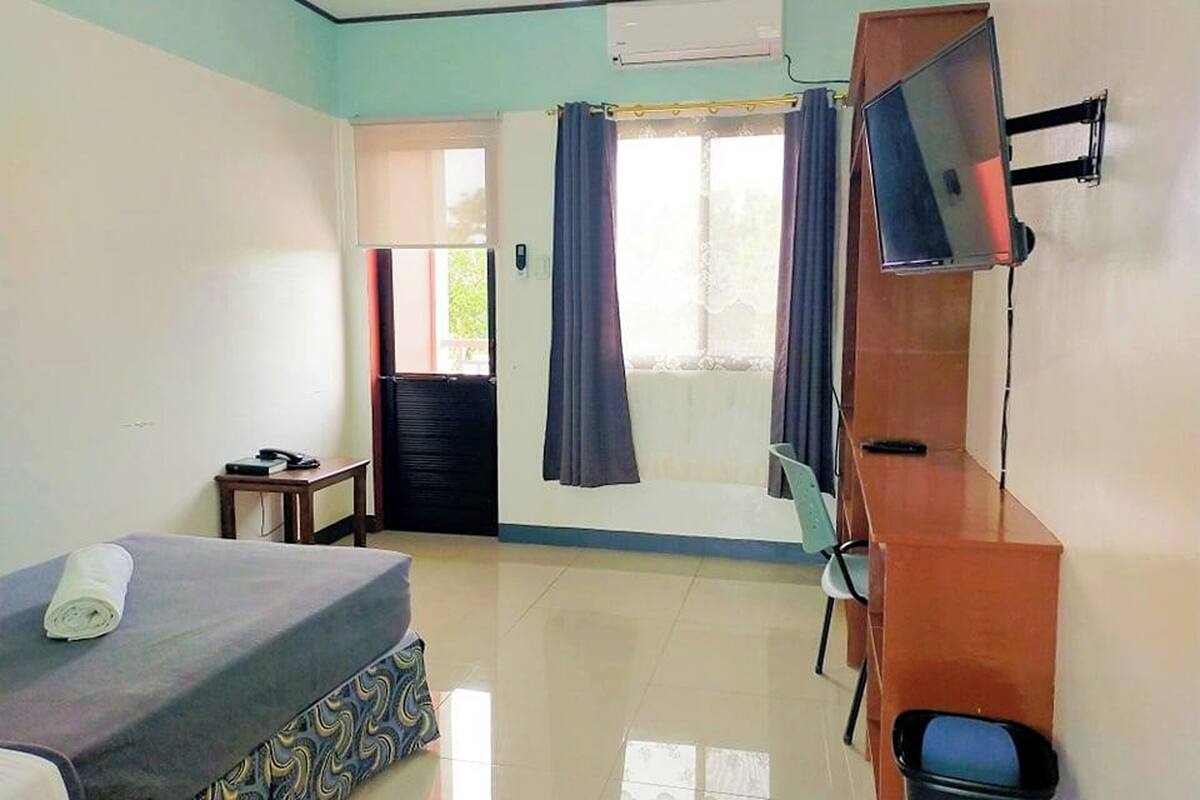 A hotel room with a bed, a desk, a chair, a TV mounted on the wall, and a window with curtains.